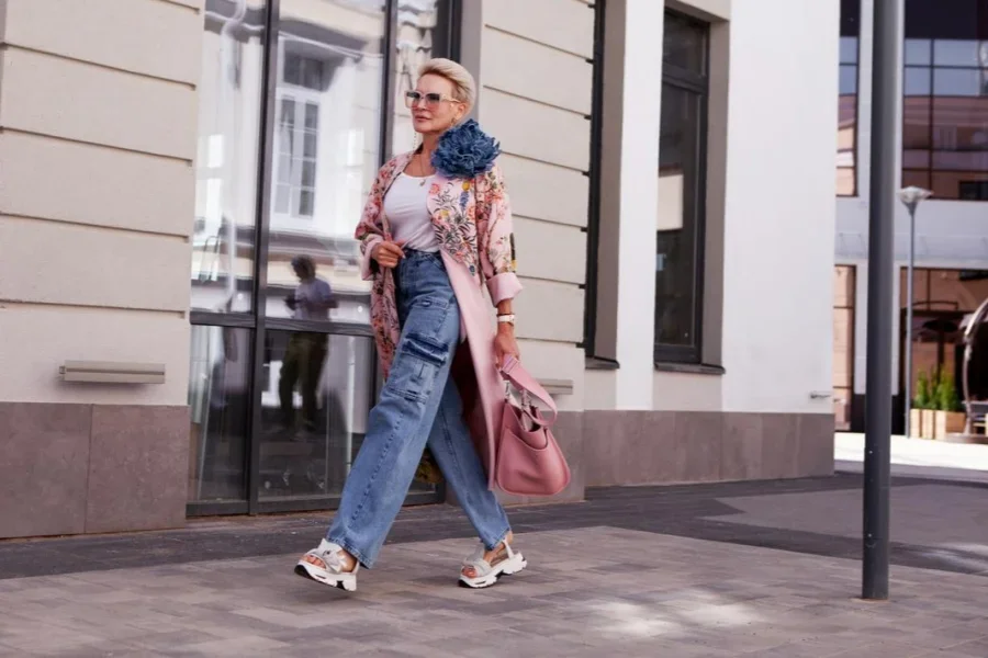 Stylish mature woman walks near city building, wearing cargo jeans, pink floral trench coat, sunglasses and handbag